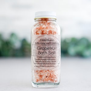 Op de afbeelding: Een heldere glazen fles gevuld met roze grapefruit badzout. De fles heeft een witte dop en een etiket met de tekst "Little Flower Grapefruit Bath Salt". De achtergrond is wazig met groen.