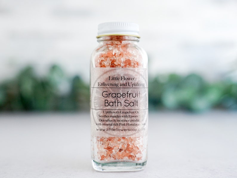 Op de afbeelding: Een heldere glazen fles gevuld met roze grapefruit badzout. De fles heeft een witte dop en een etiket met de tekst "Little Flower Grapefruit Bath Salt". De achtergrond is wazig met groen.