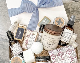 May include: A gift box with a blue striped ribbon containing a lavender soy wax candle, rose geranium face and body oil, lavender and bergamot room spray, various balms, a sugar scrub, a bath bomb, soap bars, and a small wooden spoon.