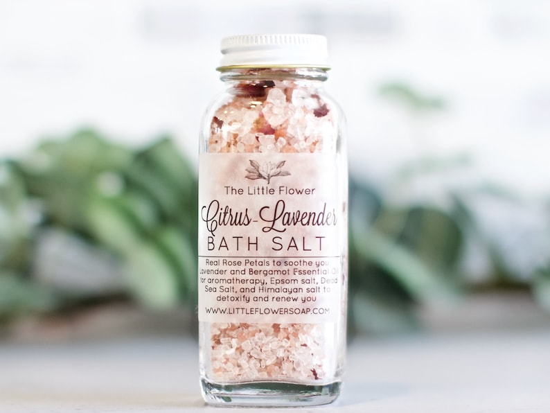Op de afbeelding: Een heldere glazen fles gevuld met roze badzout en rozenblaadjes. Het etiket leest "Citrus-Lavender BATH SALT" van "The Little Flower". De fles heeft een zilveren deksel, tegen een vage groene achtergrond.
