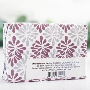 May include: A white box with a purple and silver floral pattern. The box contains a bar of soap. The ingredients are listed on the back of the box: Water, Coconut Oil, Olive Oil, Tallow, Sodium Hydroxide, Lavender Essential Oil, Lemongrass Essential Oil, Organic Lavender flowers. The website address is www.littleflowersoap.com.