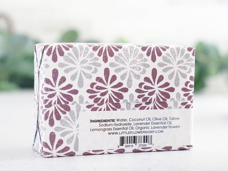 May include: A white box with a purple and silver floral pattern. The box contains a bar of soap. The ingredients are listed on the back of the box: Water, Coconut Oil, Olive Oil, Tallow, Sodium Hydroxide, Lavender Essential Oil, Lemongrass Essential Oil, Organic Lavender flowers. The website address is www.littleflowersoap.com.