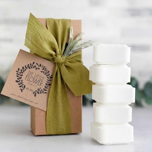 May include: A rectangular brown cardboard box wrapped in a wide olive green ribbon with a sprig of dried greenery tucked in. A square kraft paper tag features a wreath graphic and the text THE LITTLE FLOWER SOAP CO. EST. 2018. A stack of five white rectangular soap bars sits beside the box.