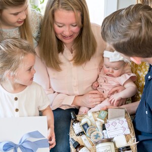 K&ouml;nnte beinhalten: Eine Familie betrachtet einen Geschenkkorb mit Pflegeprodukten. Der Korb enth&auml;lt Kerzen, Sprays, Seifen und andere Artikel. Ein Kind h&auml;lt eine wei&szlig;e Geschenkbox mit blauem Band. Die Szene wird von nat&uuml;rlichem Licht beleuchtet.
