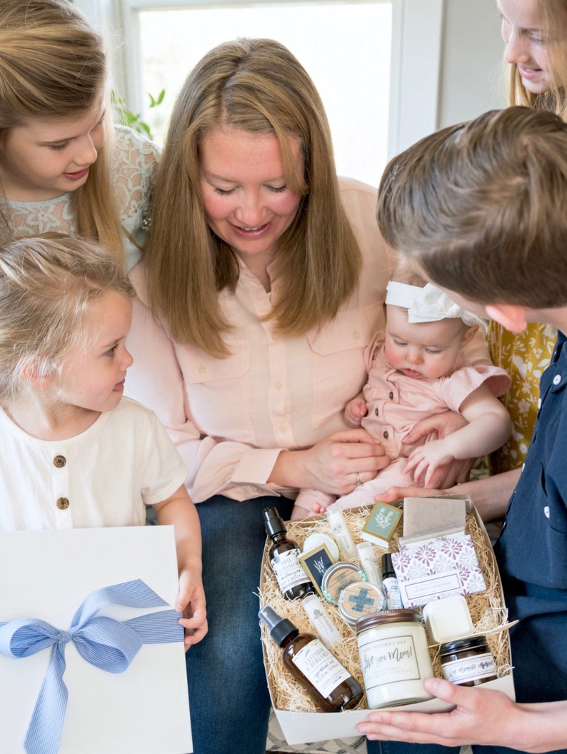 K&ouml;nnte beinhalten: Eine Familie betrachtet einen Geschenkkorb mit Pflegeprodukten. Der Korb enth&auml;lt Kerzen, Sprays, Seifen und andere Artikel. Ein Kind h&auml;lt eine wei&szlig;e Geschenkbox mit blauem Band. Die Szene wird von nat&uuml;rlichem Licht beleuchtet.