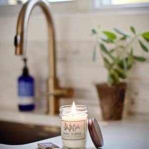 May include: A lit soy wax candle in a clear glass jar with a brown lid. The candle is labeled "Lemon Verbena Soy Wax Candle".