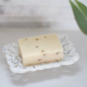 May include: A white bar of soap with flecks of herbs in a white ceramic soap dish.