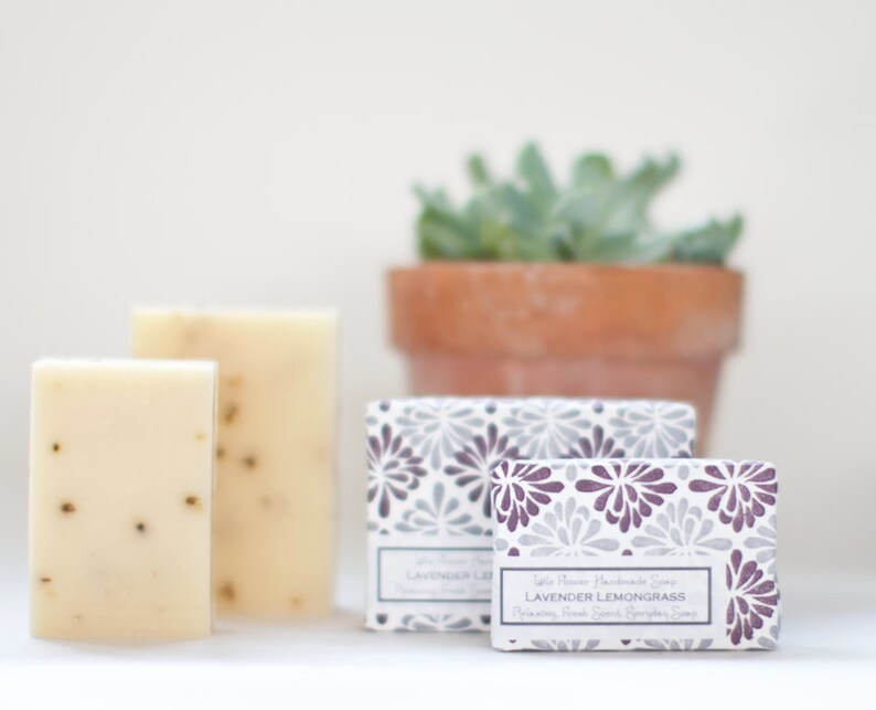 May include: Two bars of white handmade soap with lavender and lemongrass scent. The soap is packaged in a box with a purple and gray floral pattern. The text on the box reads "Little Flower Handmade Soap Lavender Lemongrass Relaxing, Fresh Scent Lemongrass Soap".