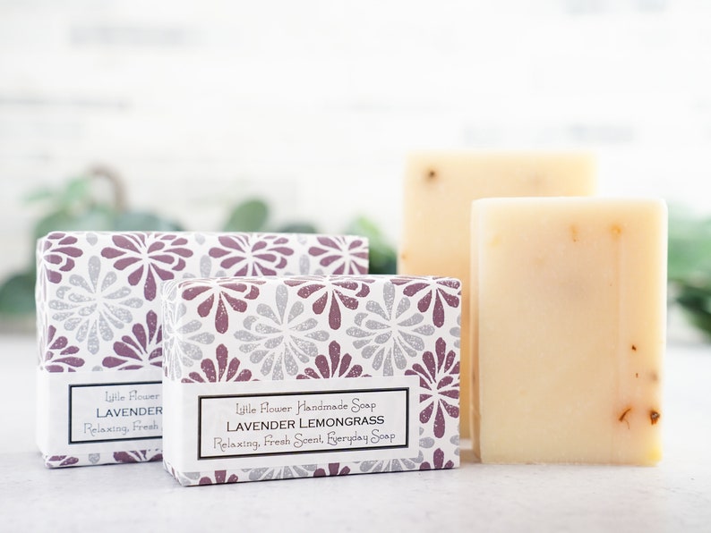 May include: Two bars of handmade soap in boxes with floral patterns. The soap is white with dried herbs and the boxes have the text "Little Flower Handmade Soap Lavender Lemongrass Relaxing, Fresh Scent, Everyday Soap" and "Little Flower Lavender Relaxing, Fresh".