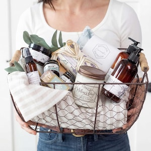 Puede incluir: Una cesta de alambre llena de productos de cuidado personal, que incluyen una vela, jab&oacute;n y un pulverizador. La cesta contiene una variedad de art&iacute;culos con etiquetas y est&aacute; decorada con vegetaci&oacute;n y una tela a rayas. El tarro de la vela dice "Happy Mother's Day".