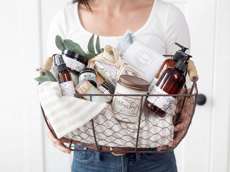 Puede incluir: Una cesta de alambre llena de productos de cuidado personal, que incluyen una vela, jab&oacute;n y un pulverizador. La cesta contiene una variedad de art&iacute;culos con etiquetas y est&aacute; decorada con vegetaci&oacute;n y una tela a rayas. El tarro de la vela dice "Happy Mother's Day".