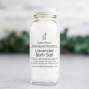 Op de afbeelding: Een heldere glazen fles gevuld met witte badzouten. Het etiket luidt "Little Flower Lavender Bath Salt" met details over lavendel etherische olie, Epsom zout en Dode Zeezout. De fles heeft een witte deksel en een gouden rand.