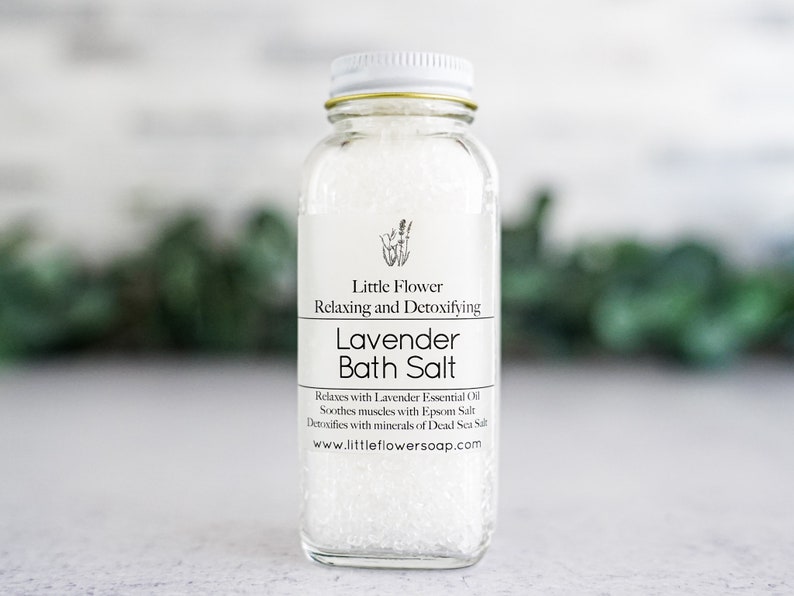 Op de afbeelding: Een heldere glazen fles gevuld met witte badzouten. Het etiket luidt "Little Flower Lavender Bath Salt" met details over lavendel etherische olie, Epsom zout en Dode Zeezout. De fles heeft een witte deksel en een gouden rand.