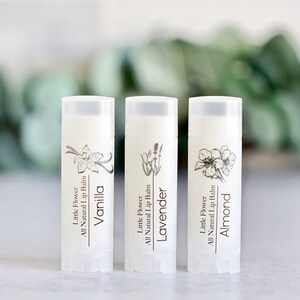 May include: Three white lip balm tubes with the text "Vanilla", "Lavender", and "Almond". Each tube has a botanical illustration and the words "Little Flower All Natural Lip Balm". The background is blurred.