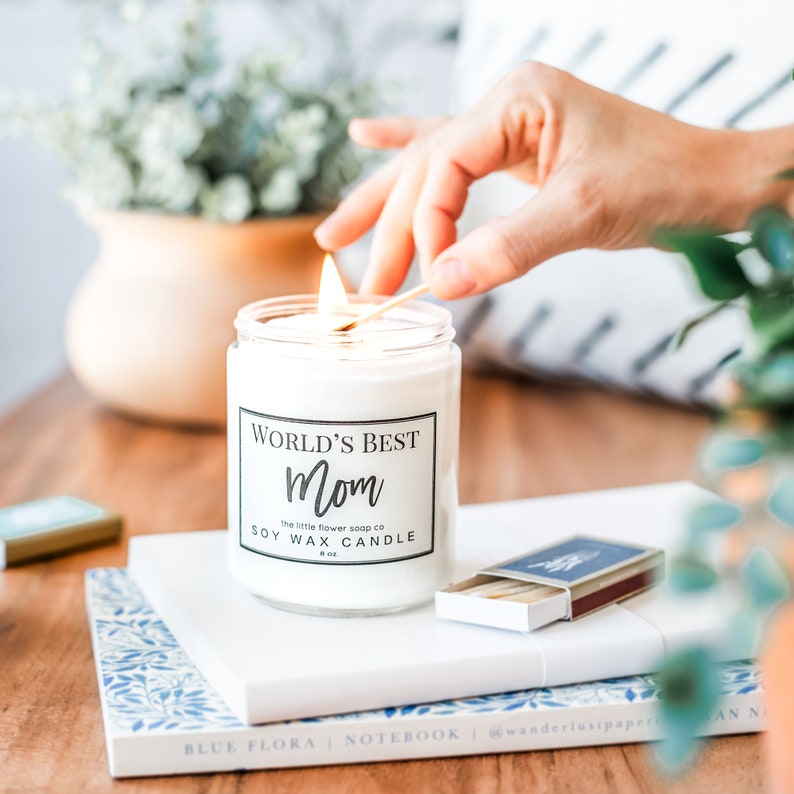 K&ouml;nnte beinhalten: Eine brennende Sojawachskerze in einem durchsichtigen Glas mit dem Text "World's Best Mom". Eine Hand z&uuml;ndet die Kerze mit einem Streichholz an. Die Kerze steht auf einem Stapel B&uuml;cher, mit einer Streichholzschachtel und einer Vase mit Gr&uuml;nzeug im Hintergrund.