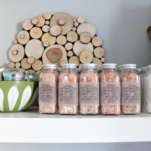 Op de afbeelding: Een display van badproducten op een witte plank. Doorzichtige glazen potten gevuld met roze grapefruit badzout en witte lavendel badzout zijn in rijen gerangschikt. Een groene kom met witte bloemmotieven bevat kleine metalen blikjes. Een houten kunstwerk hangt aan de muur.