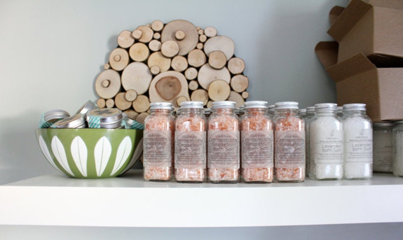 Op de afbeelding: Een display van badproducten op een witte plank. Doorzichtige glazen potten gevuld met roze grapefruit badzout en witte lavendel badzout zijn in rijen gerangschikt. Een groene kom met witte bloemmotieven bevat kleine metalen blikjes. Een houten kunstwerk hangt aan de muur.