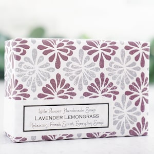 May include: A white box with a purple and silver floral pattern. The box contains a bar of lavender lemongrass soap. The label reads "Little Flower Handmade Soap Lavender Lemongrass Relaxing, Fresh Scent, Everyday Soap".