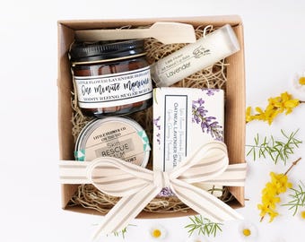 May include: A gift box filled with lavender-scented bath and body products. The box contains a jar of sugar scrub labeled "One Minute Manicure", a small tin of skin balm labeled "Skin Rescue Balm", a bar of soap labeled "Oatmeal Lavender Sage", and a tube of lip balm labeled "Lavender".