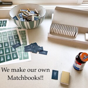 May include: A flat lay image of a table with various items, including a bowl filled with matchbooks, a box of matchbooks, a stack of printed cards, a glue bottle, and a matchbook with the text "We make our own Matchbooks!!".