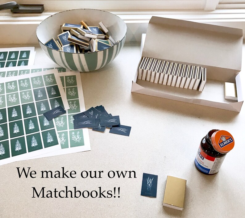 May include: A flat lay image of a table with various items, including a bowl filled with matchbooks, a box of matchbooks, a stack of printed cards, a glue bottle, and a matchbook with the text "We make our own Matchbooks!!".
