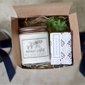 May include: A gift box with a white soy wax candle with a brown lid and a label that reads "Balance & Uplift Soy Wax Candle" and a bar of soap with a white and gray floral pattern and the text "Lavender Lemongrass" on the label.