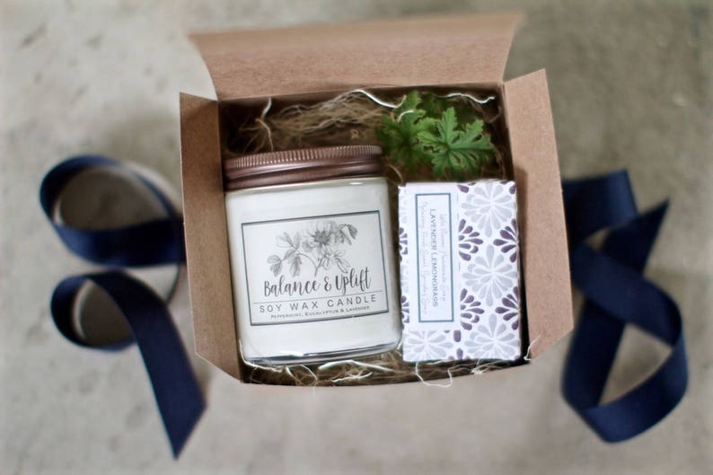 May include: A gift box with a white soy wax candle with a brown lid and a label that reads "Balance & Uplift Soy Wax Candle" and a bar of soap with a white and gray floral pattern and the text "Lavender Lemongrass" on the label.