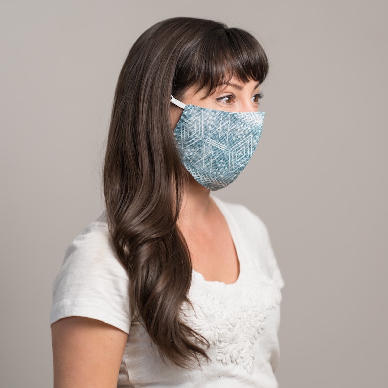 May include: A woman with long brown hair is wearing a blue and white patterned face mask. The mask has a geometric design and is covering her nose and mouth.