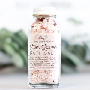 May include: A glass jar filled with pink bath salts and rose petals. The label reads "The Little Flower Citrus-Lavender Bath Salt" and includes information about the ingredients and website.