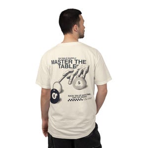 Könnte beinhalten: Cremefarbenes T-Shirt mit einer Schwarz-Weiß-Grafik einer Hand, die einen Billardqueue positioniert, um die Kugel mit der Nummer 5 zu treffen. Der Text "MASTER THE TABLE" und "RACK 'EM UP ANYTIME, DAY OR NIGHT" sind über und unter der Grafik gedruckt.