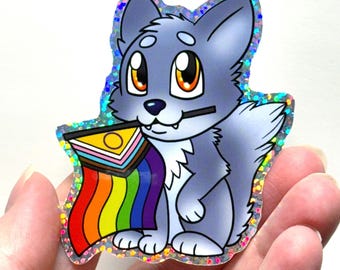 May include: A holographic sticker featuring a cartoon wolf with gray fur and large orange eyes. The wolf is holding a rainbow pride flag in its mouth. The sticker has a glittery, iridescent border.