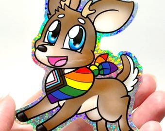 May include: A holographic sticker featuring a cartoon deer with large blue eyes and a brown coat. The deer wears a colorful bandana displaying the rainbow pride flag and the progress pride flag colors. The sticker has a shimmering finish.