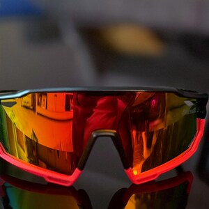 May include: Sport sunglasses with a black frame and vibrant, mirrored lenses reflecting a spectrum of colors. The lower frame is outlined in bright red. These sunglasses are designed for outdoor activities.