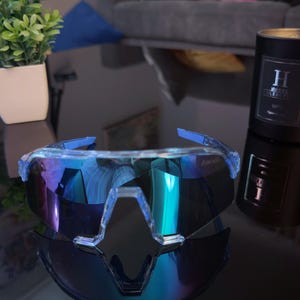 May include: Sport sunglasses with a clear frame and blue accents. The lenses have a mirrored, iridescent finish with shades of blue, purple, and green. A black candle with white text is also visible.