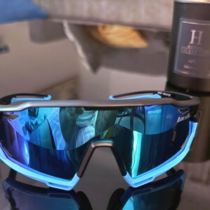 May include: Sport sunglasses with a black frame and blue mirrored lenses. The sunglasses have a light blue accent on the frame. The lenses reflect the surrounding environment. The sunglasses are on a reflective surface.