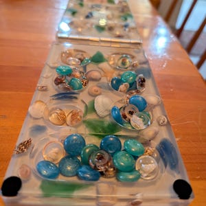 May include: A rectangular resin tray filled with seashells, blue and teal glass beads, and green glass pieces. The tray has a clear base with a light blue border and black corner accents. The tray is on a wooden table.