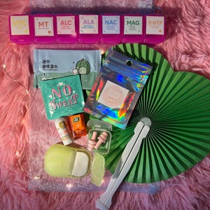 Może przedstawiać: Zestaw produktów zdrowotnych i wellness, w tym różowy organizer na pigułki z etykietami suplementów, zielony wachlarz oraz małe opakowania chusteczek i innych produktów. Tekst "NO SWEAT" jest widoczny na turkusowym opakowaniu.