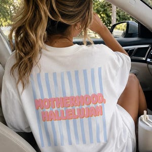 May include: White t-shirt featuring the words "MOTHERHOOD, HALLELUJAH" in pink and orange, with light blue vertical stripes. The shirt is worn by a person in a car.