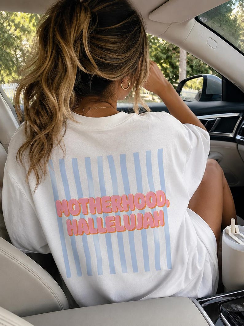 May include: White t-shirt featuring the words "MOTHERHOOD, HALLELUJAH" in pink and orange, with light blue vertical stripes. The shirt is worn by a person in a car.
