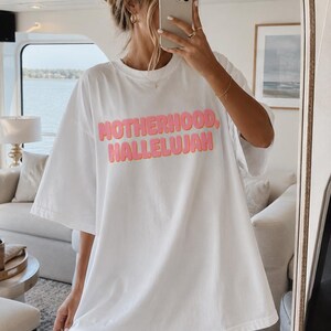 May include: White oversized t-shirt with the words "MOTHERHOOD, HALLELUJAH" in pink, bubble letters. The shirt is being worn by a person in front of a mirror. The person is holding a phone up to their face.