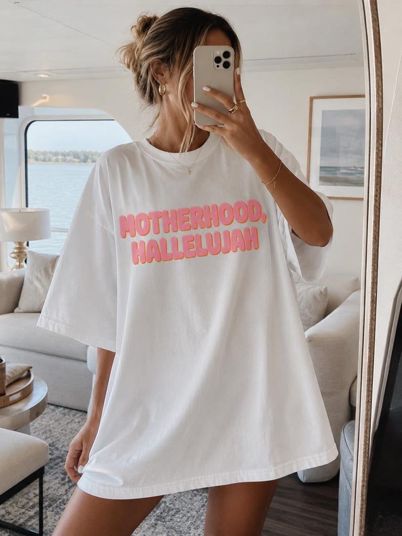 May include: White oversized t-shirt with the words "MOTHERHOOD, HALLELUJAH" in pink, bubble letters. The shirt is being worn by a person in front of a mirror. The person is holding a phone up to their face.