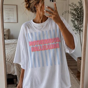 May include: White oversized t-shirt with the words "MOTHERHOOD, HALLELUJAH" in pink and blue. The shirt features vertical light blue stripes. A casual, comfortable style.