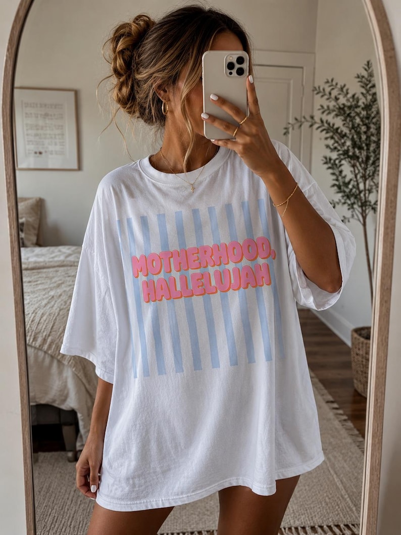 May include: White oversized t-shirt with the words "MOTHERHOOD, HALLELUJAH" in pink and blue. The shirt features vertical light blue stripes. A casual, comfortable style.