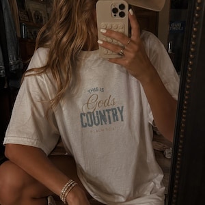以下が含まれることがあります： 青い文字で「THIS IS GOD'S COUNTRY」と書かれた白いTシャツ。Tシャツには「PSALM 50:11」の文字も含まれています。カウボーイハットをかぶり、携帯電話を持っています。