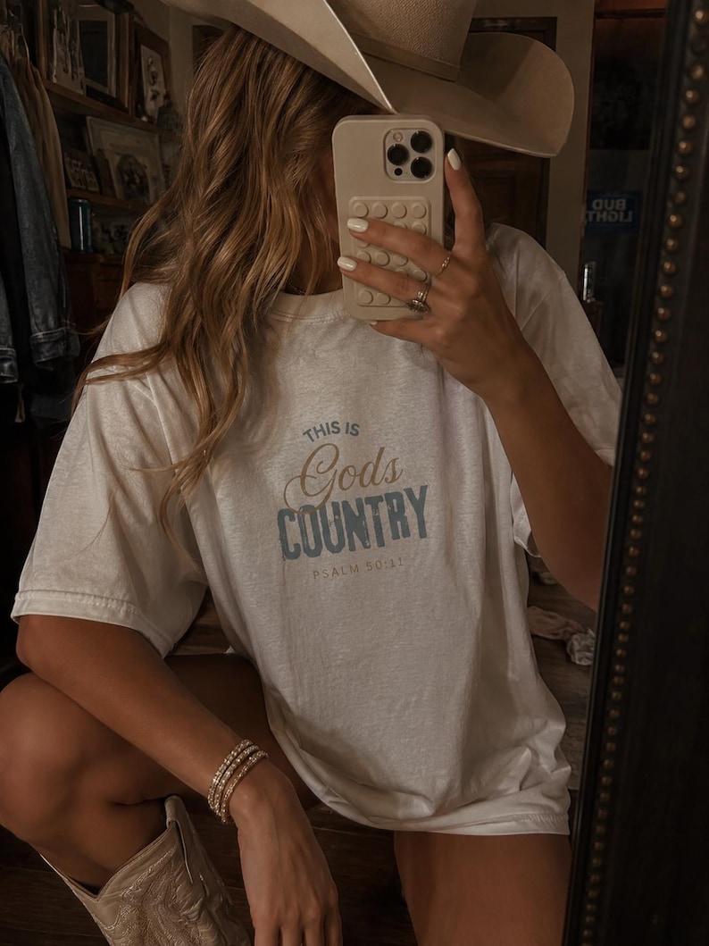 以下が含まれることがあります： 青い文字で「THIS IS GOD'S COUNTRY」と書かれた白いTシャツ。Tシャツには「PSALM 50:11」の文字も含まれています。カウボーイハットをかぶり、携帯電話を持っています。