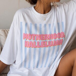 May include: White t-shirt featuring the text "MOTHERHOOD, HALLELUJAH" in pink bubble letters, with light blue vertical stripes. The shirt has an oversized fit and is likely made from a soft fabric.