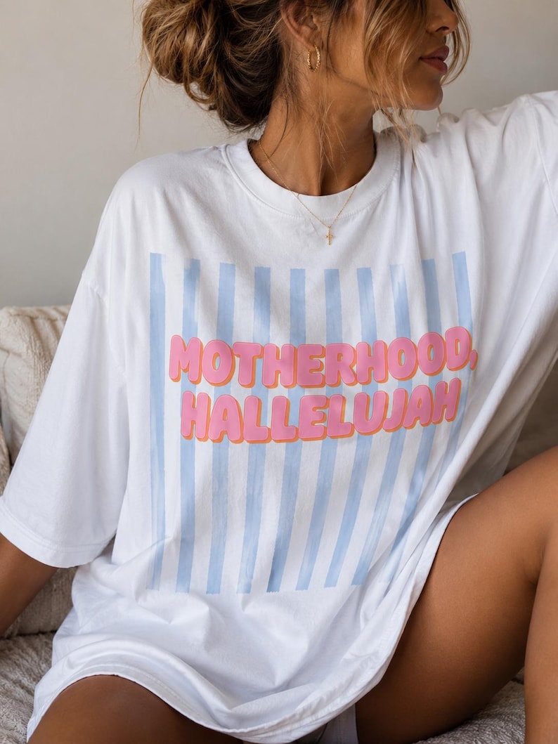 May include: White t-shirt featuring the text "MOTHERHOOD, HALLELUJAH" in pink bubble letters, with light blue vertical stripes. The shirt has an oversized fit and is likely made from a soft fabric.