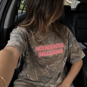 May include: A camouflage-patterned t-shirt with the words "MOTHERHOOD, HALLELUJAH" in pink block letters. The shirt is worn with grey sweatpants. The image is taken from inside a car.