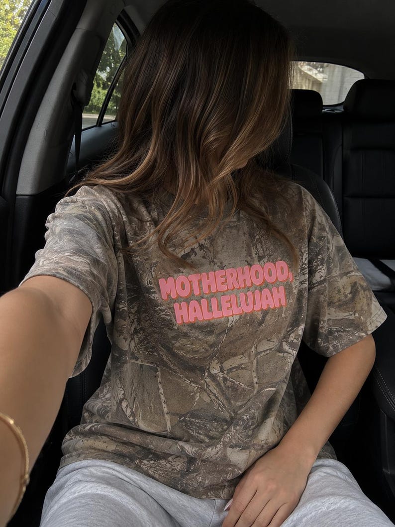 May include: A camouflage-patterned t-shirt with the words "MOTHERHOOD, HALLELUJAH" in pink block letters. The shirt is worn with grey sweatpants. The image is taken from inside a car.
