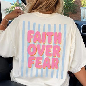 May include: Cream-colored t-shirt with the words "FAITH OVER FEAR" in pink bubble letters with orange outlines. The text is set against a backdrop of vertical light blue stripes. The shirt is a casual, everyday wear item.